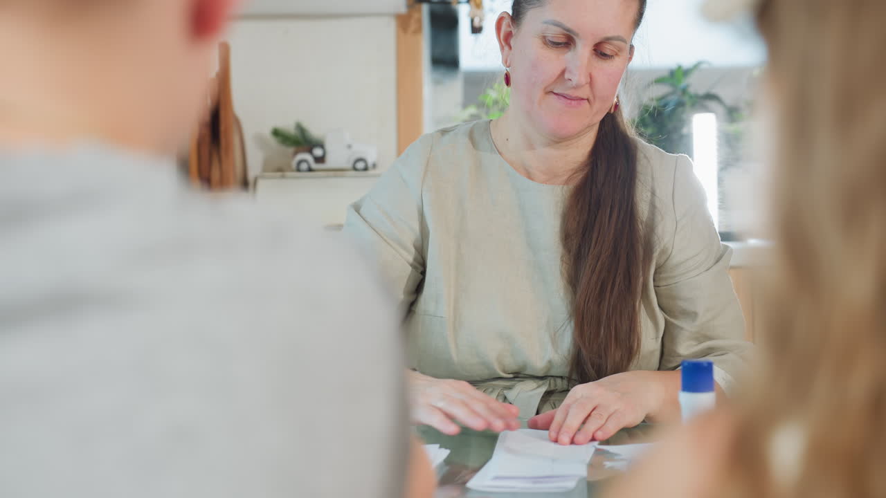 Woman in gray gown sitting folding paper on table with two kids in front blurred view indoor home environment, focus on paper folding activity with warm atmosphere and calm setting