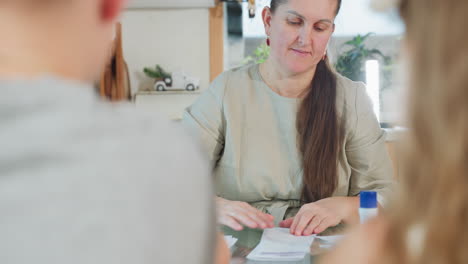 woman in gray gown sitting folding paper on table with two kids in front blurred view indoor home environment, focus on paper folding activity with warm atmosphere and calm setting