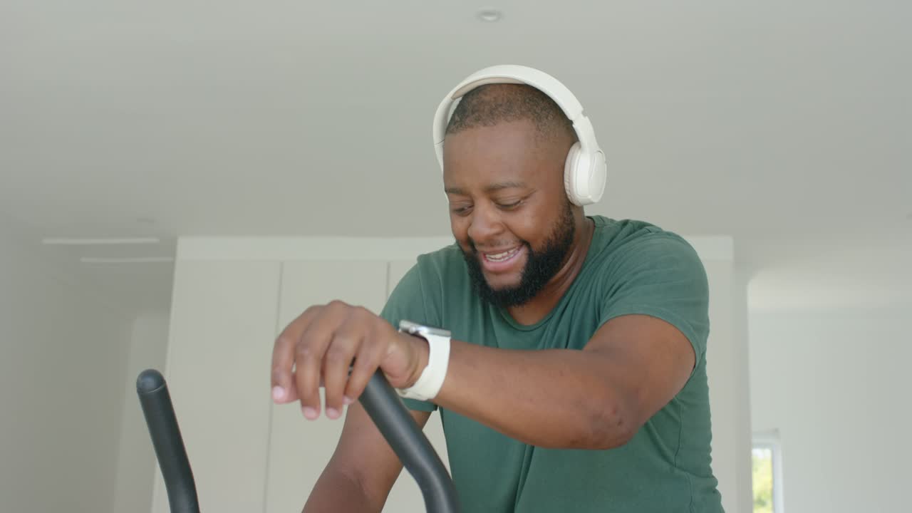 African American man in green tee using console pedaling bike at home checking smartwatch for stats