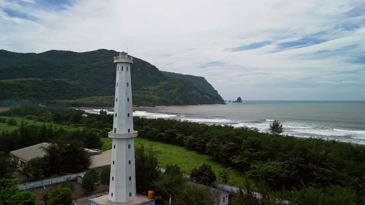 Video flycam trên không cho thấy một ngọn hải đăng trắng cao đứng trên sườn đồi ven biển xanh mướt, với tầm nhìn ra đại dương rộng lớn, sóng biển êm dịu và môi trường bờ biển tự nhiên yên bình dưới ánh sáng ban ngày dịu nhẹ.