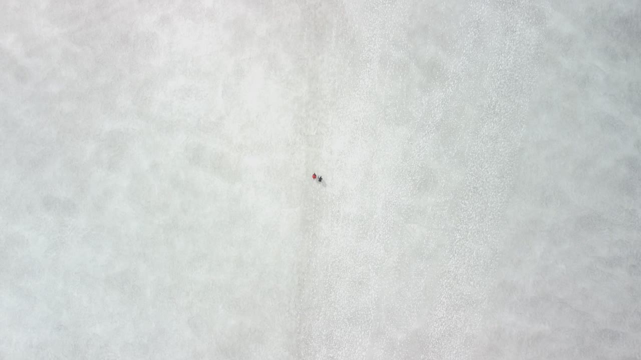 Top down aerial view of two hikers walking on white snow and ice covered landscapes in Iceland during a clludy day. People are walking forward and drone moves with them with smooth motion.
