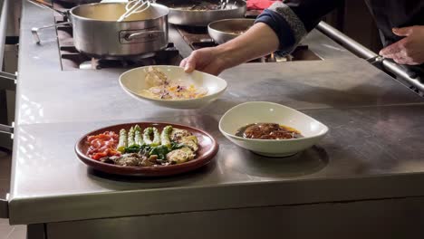 chef wearing black dress places red fried mullet by a asparagus and roasted pepper salad and a foie gras with sherry and brandy sauce, over the professional restaurant's kitchen counter