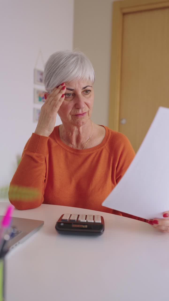 Donna anziana che calcola le finanze a casa con laptop e documenti.