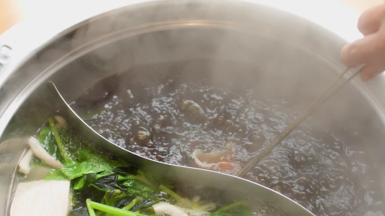 Chopsticks cook sliced meat in steaming dual broth hotpot with fresh vegetables, overhead view