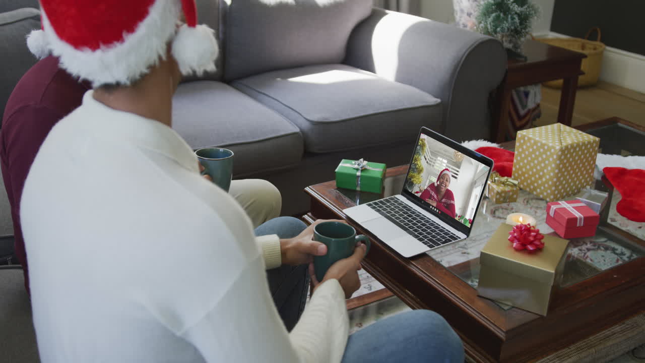 padre y hijo biraciales usando una computadora portátil para una videollamada de navidad con una mujer feliz en la pantalla
