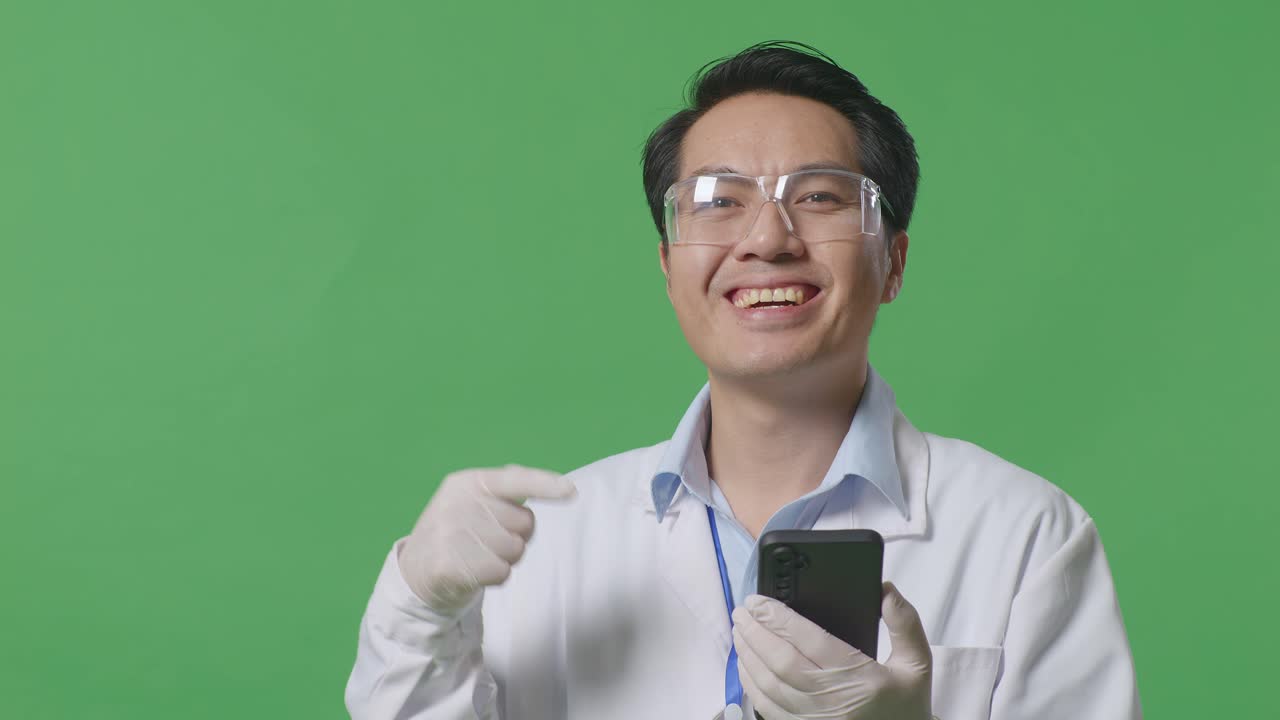 아시아인 과학자가 미소 짓고 스마트폰을 가리키며 실험실의 녹색 화면 배경에 서 있습니다.