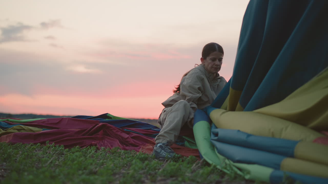 woman guiding deflated hot air balloon canopy into basket on green field during sunset landing sequence with pastel sky horizon showing calm rural landscape and gentle breeze