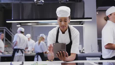 african american female chef wearing chefs whites in a restaurant kitchen, using a tablet