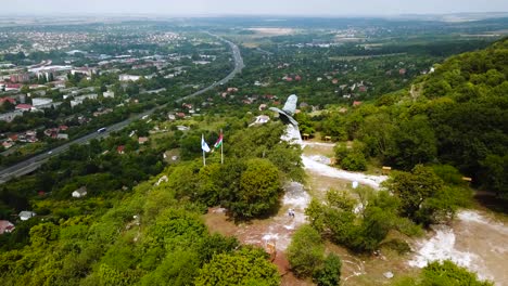 Volando-Sobre-La-Ciudad-De-Tatabánya-En-Hungría,-La-Estatua-Del-Pájaro-Turul-Y-La-Vista-De-Las-Banderas-Húngaras