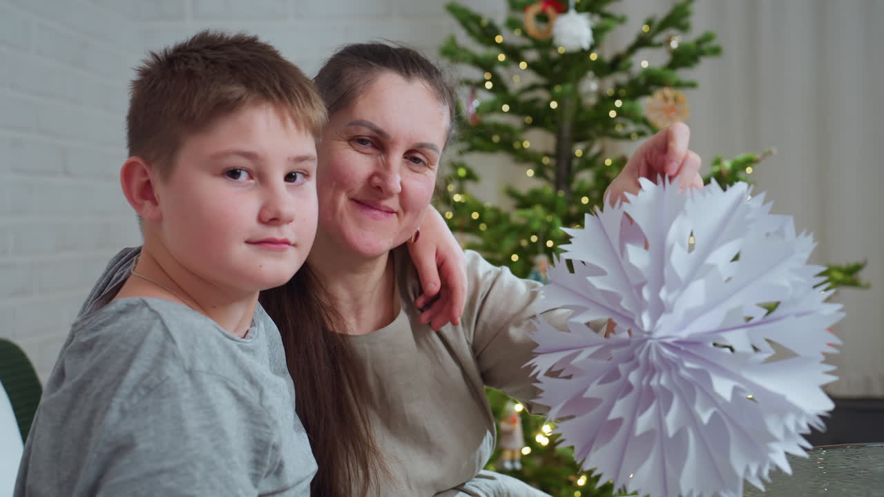 son crosses arm over mother s shoulder while mother proudly holds crafted paper snowflake near decorated christmas tree both smiling warmly and focusing on festive handmade holiday ornament indoors