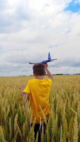 little boy plays on factory background. child in yellow t-shirt walking on wheat field and holds toy plane in his hand in summer. vertical video
