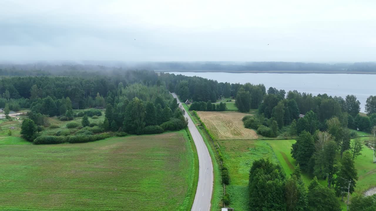 Aerial tracking shot of road by lake with misty sky, fields, and forest fading into fog