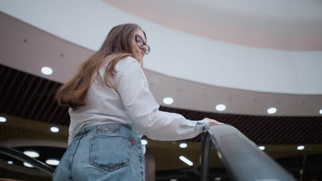 back view of young lady on moving escalator, phone in back pocket, hand on rail, looking around in modern mall with bright ceiling lights, showcasing urban lifestyle and casual shopping experience