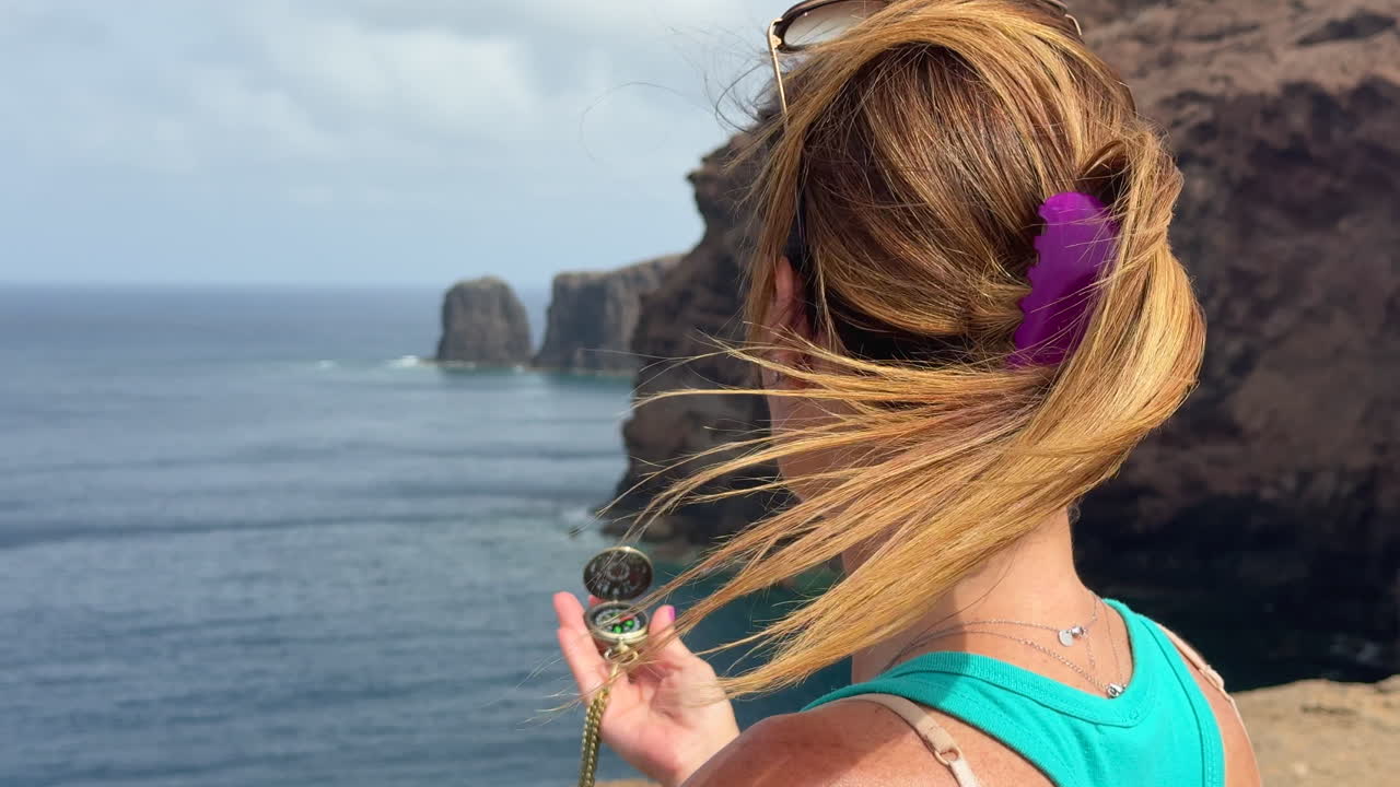 Female traveler navigating coastal terrain of Gran Canaria, holding compass with iconic Roque Partido landmark rising dramatically in rocky background during summer adventure journey
