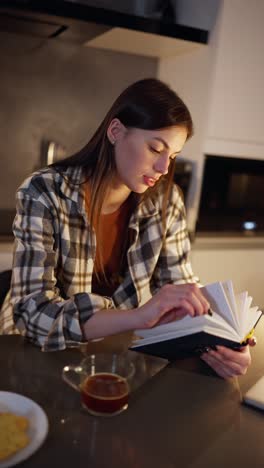 video vertical de una chica morena feliz y segura en una camisa a cuadros sentada en la mesa después de cenar y leyendo un libro en un apartamento moderno