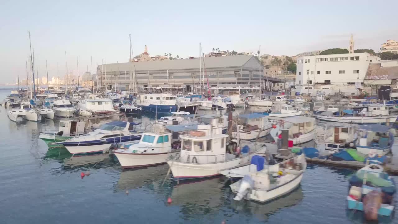 Ripresa aerea ascendente del porto e del paesaggio urbano di Jaffa con barche, sotto cieli azzurri e sereni e lo skyline di Tel Aviv in lontananza.