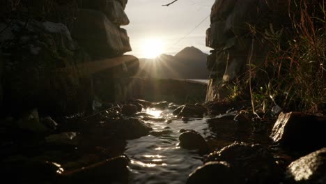 the camera slowly tilts down to reveal the sun setting behind a mountain in the background as a small river gently flows under an arch in a stone wall
