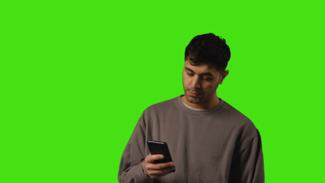 Young-Man-Looking-At-Text-Message-Or-Browsing-Online-On-Mobile-Phone-Standing-Against-Green-Screen-Background