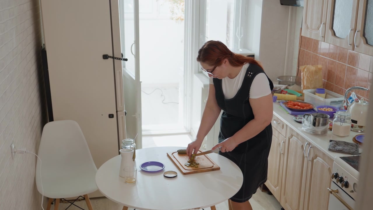 Kvinne står og lener seg over et rundt kjøkkenbord mens hun skjærer mat på et skjærebrett av tre med kniv, hender i bevegelse og pene skiver synlige, i et lyst hjemmekjøkken med bakgrunnsbelyst vindu
