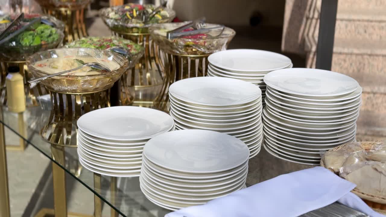 Un plano con ángulo elevado de un bufé al aire libre con hummus fresco, ensaladas mediterráneas en cuencos de cristal y pilas ordenadas de platos blancos. Perfecto para contenido de catering, bodas y hostelería