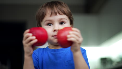 cute baby boy holding two apples in hands. kid bumps the fruit and then takes a bite. close up. blurred backdrop.