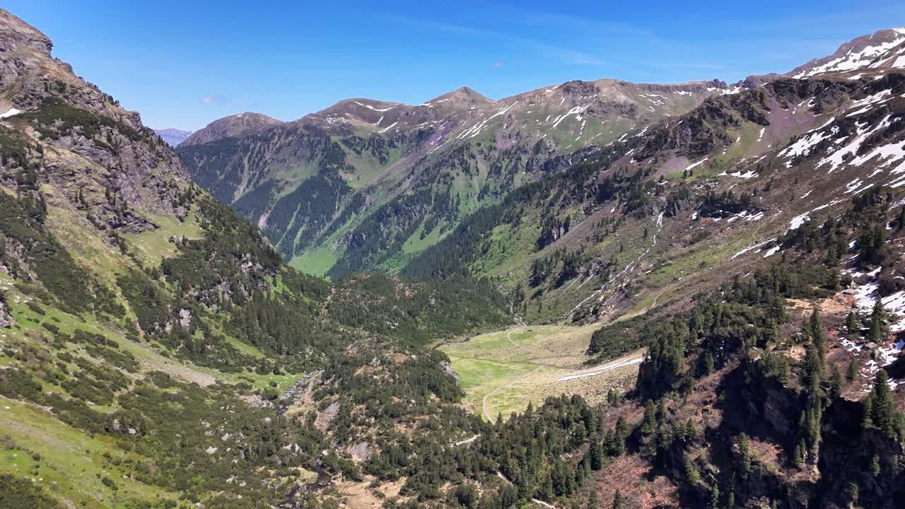 Drone pulling away from the Murgsee valley, revealing the expansive alpine landscape with lush green slopes, rocky peaks, and remnants of snow under a clear blue sky, St Gallen, Switzerland