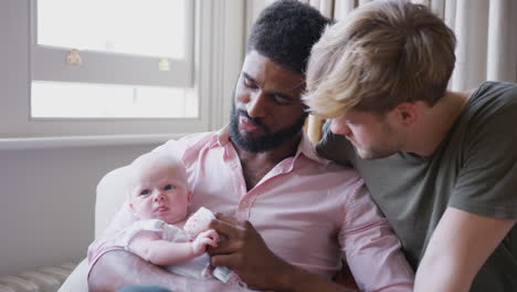 loving male same sex couple cuddling baby daughter on sofa at home together