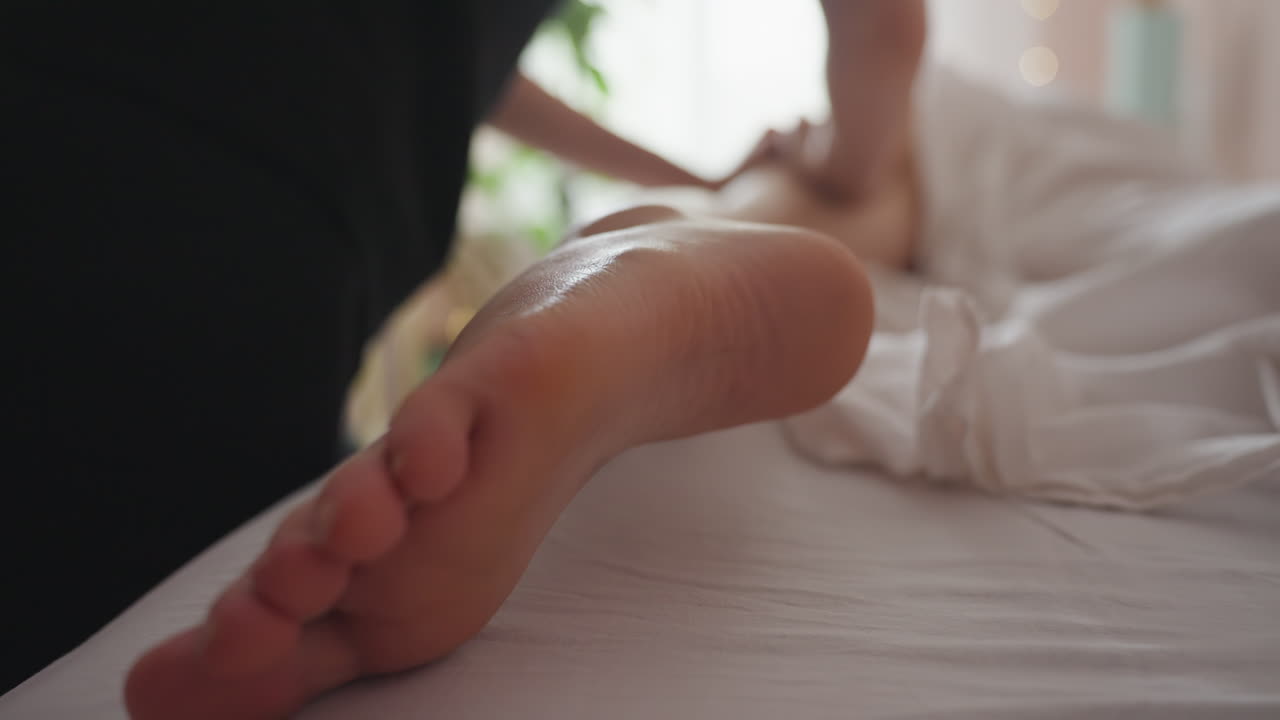 Massage therapist applies firm pressure to client leg while other leg is covered with white sheet, close-up of foot and toes visible in foreground with soft light and blurred indoor background