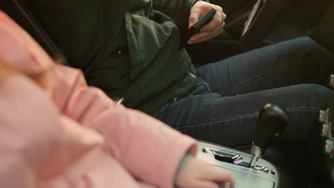 a close-up shot of a man in a green jacket seated in a car, captured as he removes his seat belt. next to him, a child in a pink jacket sits beside the center console