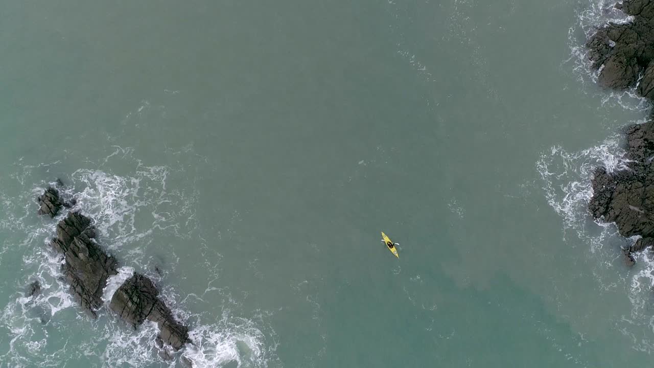 antena de arriba hacia abajo descendiendo sobre una persona en un kayak amarillo navegando alrededor de una costa rocosa en aguas turbias del mar azul