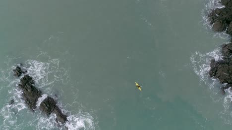 Antena-De-Arriba-Hacia-Abajo-Que-Desciende-Sobre-Una-Persona-En-Un-Kayak-Amarillo-Navegando-Alrededor-De-Una-Costa-Rocosa-En-Aguas-Turbias-Del-Mar-Azul