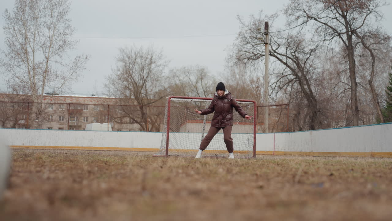 goalkeeper ready stance, ball approaches goalkeeper against overcast sky on vacant sports ground, defensive player in winter gear prepares for action beneath bleak sky on deserted urban football field