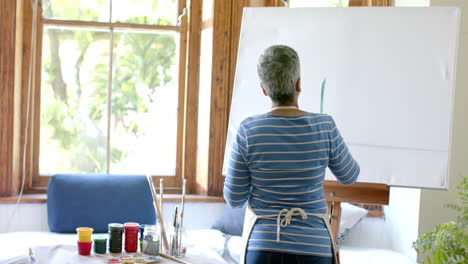 Mujer-Birracial-Mayor-Usando-Delantal-Y-Pintando-Sobre-Lienzo-Grande-En-Casa,-Cámara-Lenta