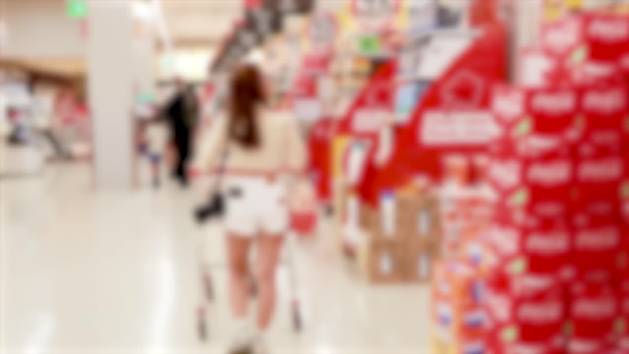A young woman in casual summer attire pushes a shopping cart down a brightly lit supermarket aisle lined with colorful product displays. Slight camera blur suggests motion