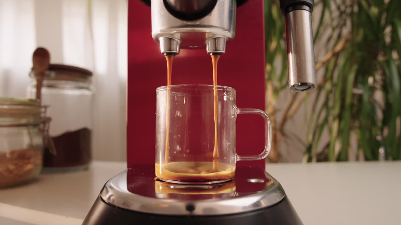 Close-up of espresso pouring into large transparent glass cup in slow motion. Crema forming as coffee fills the cup. Red coffee machine in modern home kitchen environment