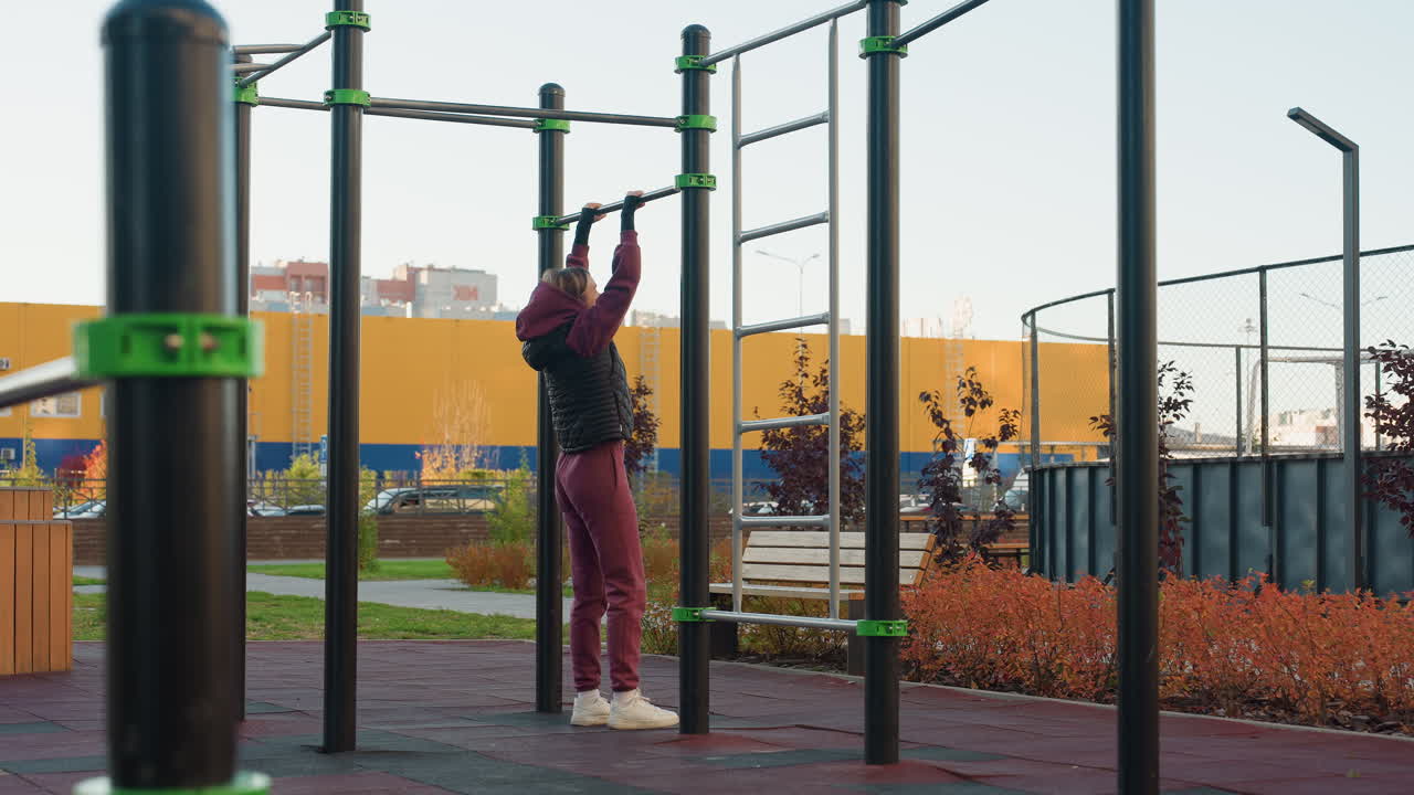 Side view female body builder pulling herself on horizontal bar in urban park fitness area on rubber flooring emphasizing forearm strength control focus during calisthenics routine
