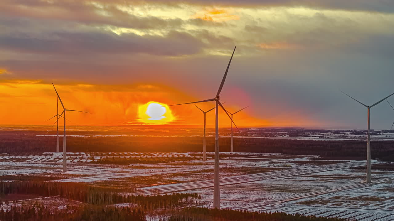 Timelapse of a winter sunset over snowy farmland with wind turbines standing still, glowing orange horizon and layered clouds creating a calm renewable energy landscape