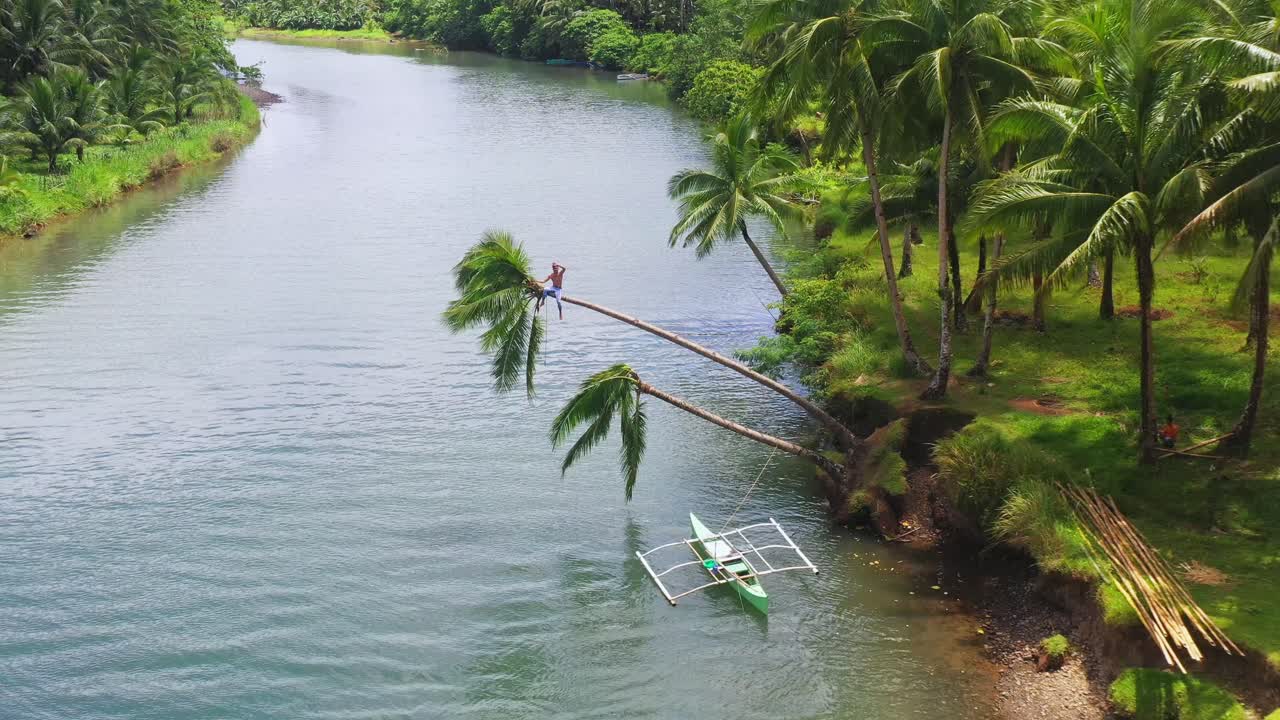 joven filipino saludando a un dron mientras se sienta en el tronco de un cocotero inclinado en la orilla del río