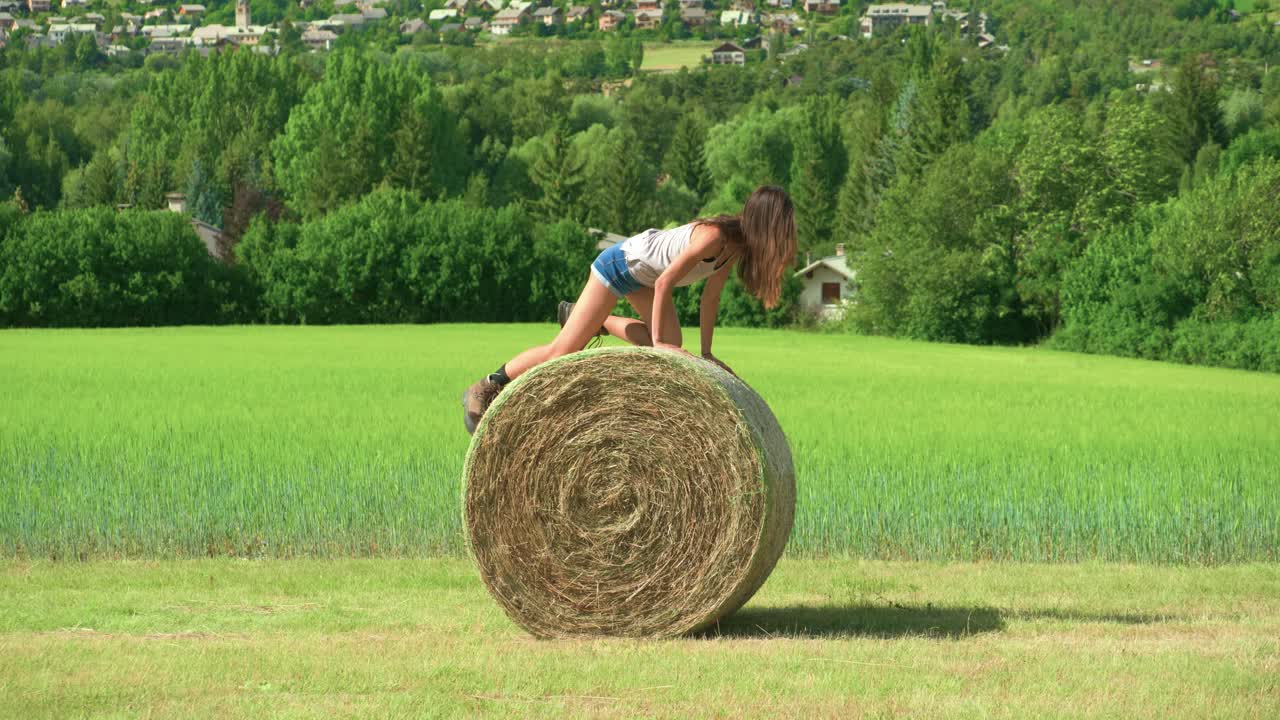 una hermosa mujer morena jugando tratando de hacer rodar una gran paca de heno en una granja en francia