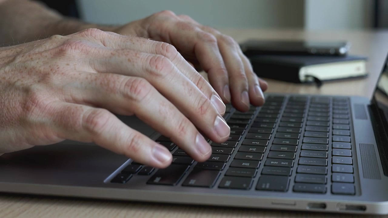 Typing adult hands pressing keys at desk, completing work with laptop, notebook nearby
