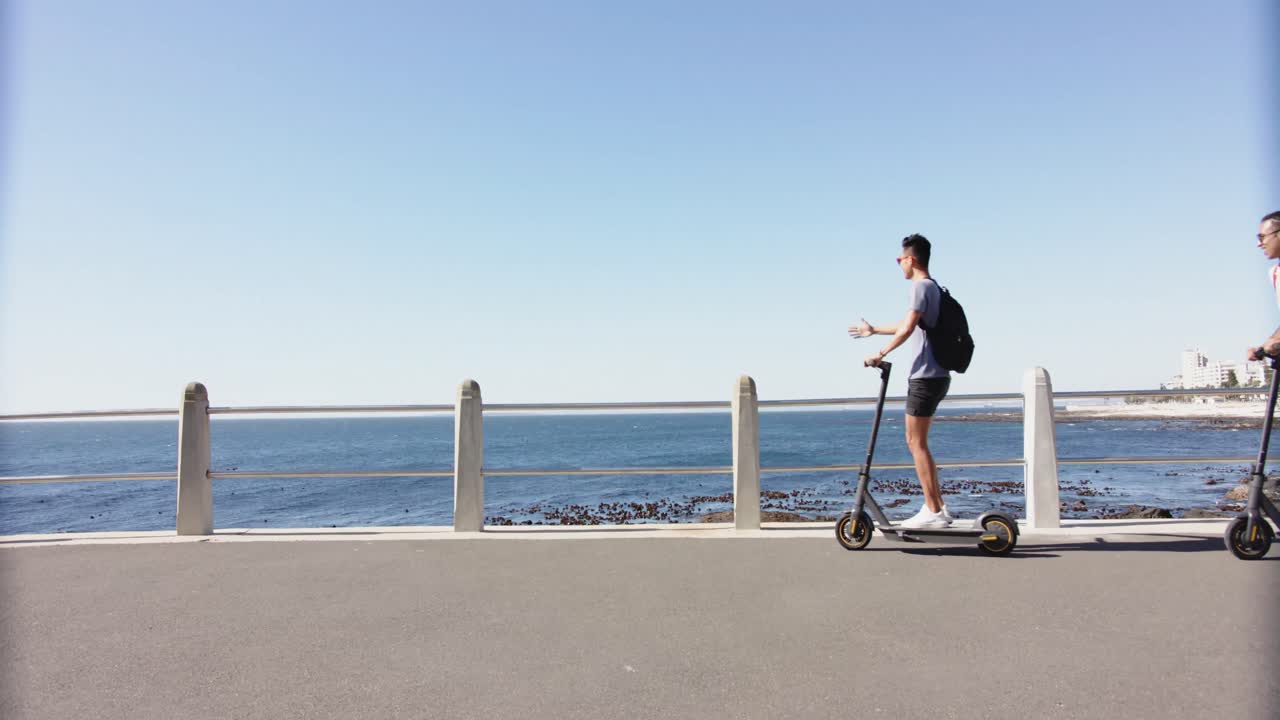 Feliz pareja gay diversa usando patinetes en el paseo marítimo, a cámara lenta.