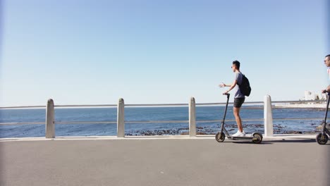 Happy-diverse-gay-male-couple-using-scooters-at-promenade-by-the-sea,-slow-motion