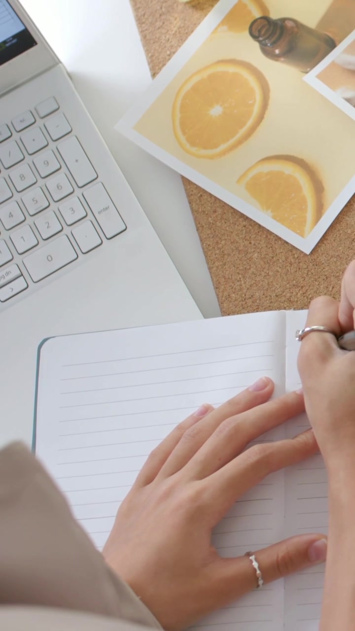 Vertical video: Lifting pen, woman right hand writing in notebook at desk, working on laptop, vials