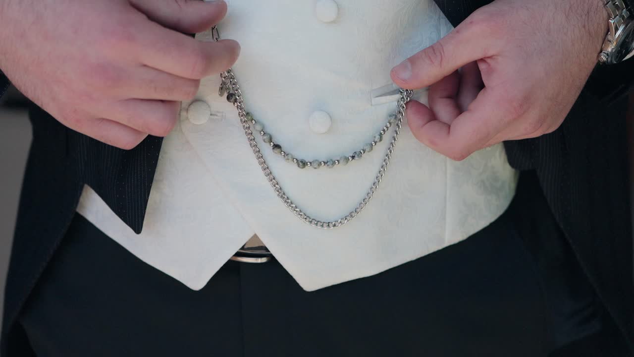 close up of groom's hands adjusting decorative chain on elegant white waistcoat