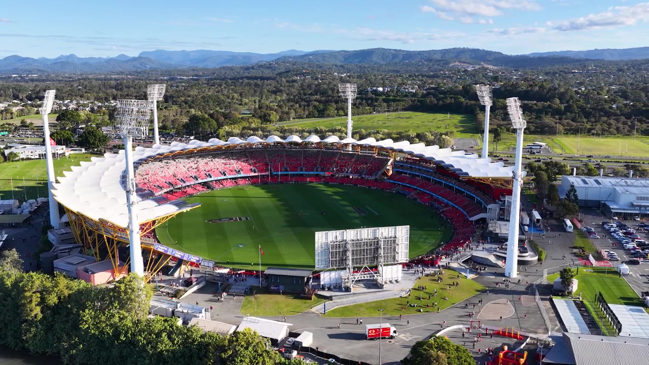 Wide aerial drone shot of a circular sports arena with green field under bright sunlight.