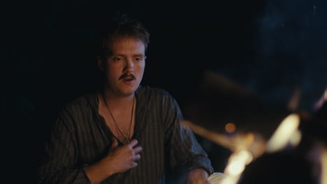 man with a mustache sitting by a campfire at night
