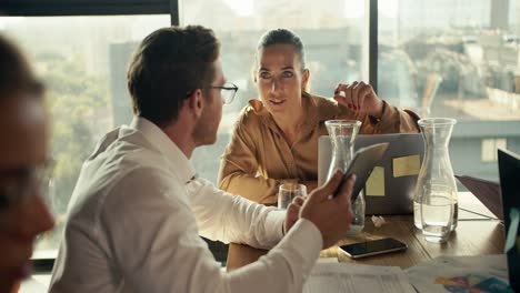 An-office-worker-in-a-shirt-and-glasses-shows-his-project-on-a-tablet-to-a-blonde-woman,-sitting-at-a-table-in-an-office-with-panoramic-windows