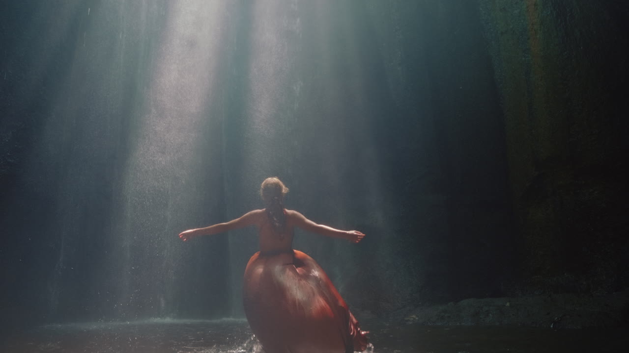 mujer feliz bailando en la cueva de la cascada salpicando agua usando un hermoso vestido disfrutando de la naturaleza bailando sintiendo libertad espiritual 4k