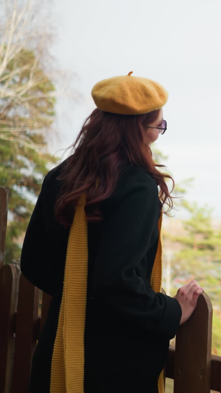 Vista laterale di una signora elegante con basco giallo e cappotto nero che si avvicina a una recinzione decorativa in legno, appoggiandovi la mano, mentre un gazebo e alberi autunnali con foglie secche sono sparsi tutt'intorno.