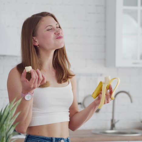 Pretty woman enjoys eating banana. Young female in casual clothing eats fresh fruit in the kitchen. Healthy food concept.